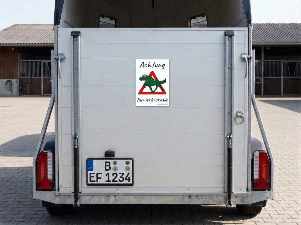 Die Rückansicht eines Pferdeanhängers. Auf der Heckklappe ist ein Schild angebracht mit einem handgezeichneten Cartoon "Achtung Turnierkrokodile".
