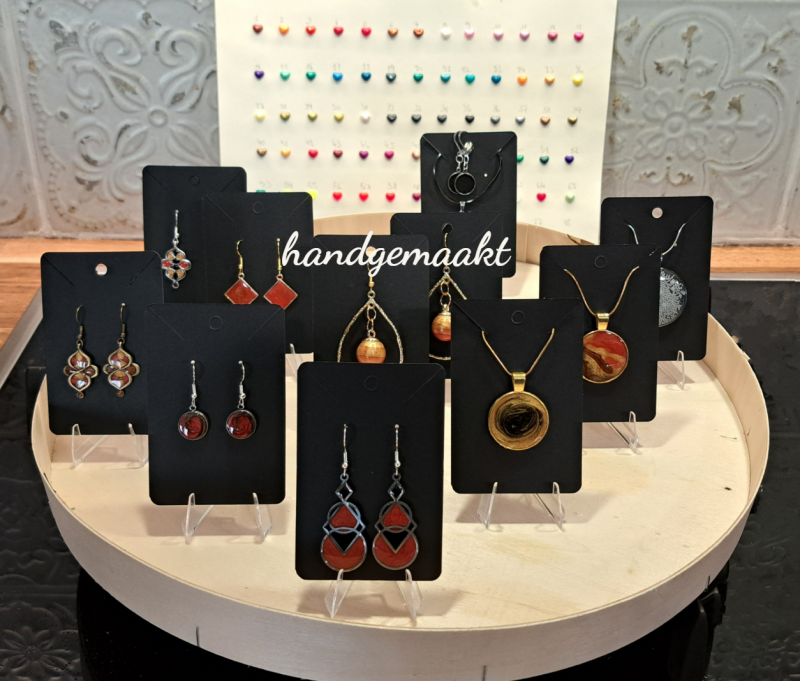 Presentatie van handgemaakte sieraden in epoxyhars, waaronder oorbellen en halskettingen, uitgestald op zwarte kaarten op een ronde houten display, met een achtergrond van kleurrijke voorbeelden.
