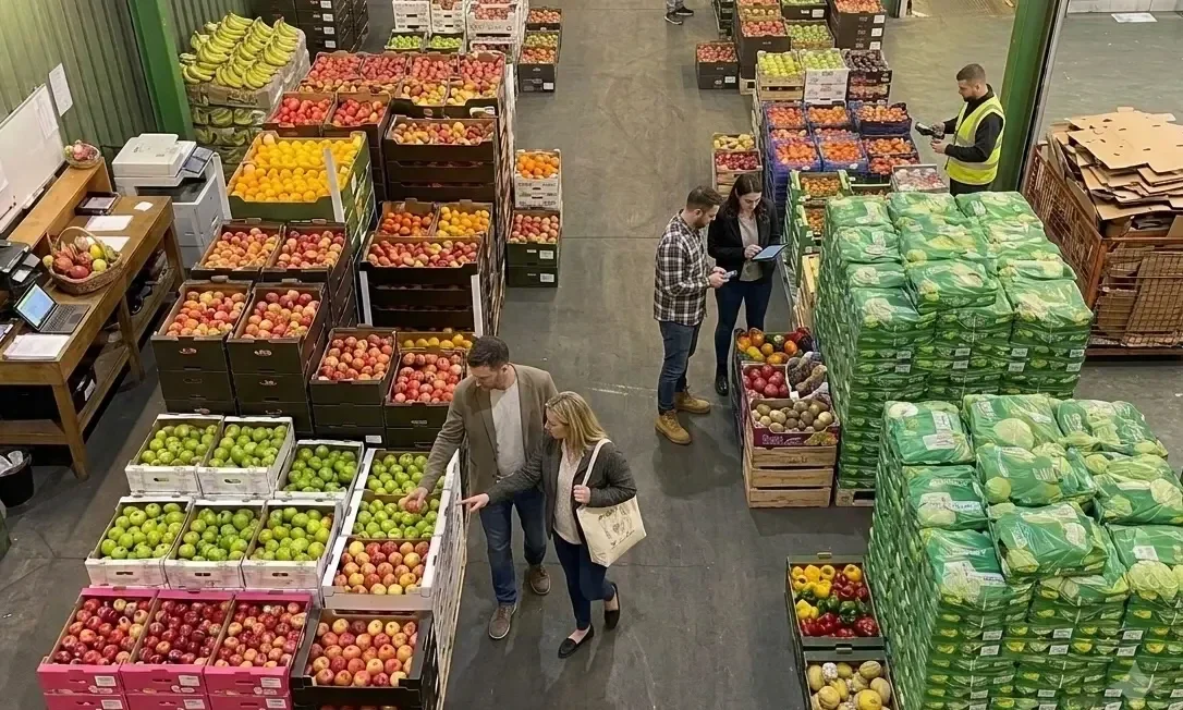 Buyers selecting fresh fruit and vegetables at Flossies Fine Foods stand in New Covent Garden Market, London, surrounded by crates of apples, oranges and bananas