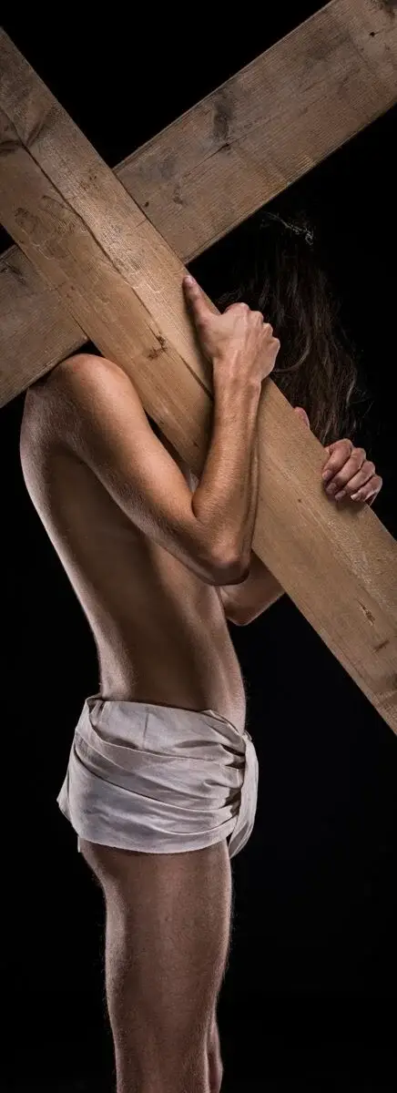 Naakte man van kleur draagt houten kruis op de schouder. Warme huidtinten en clair-obscur belichting refereren aan historische Jezus-figuur.