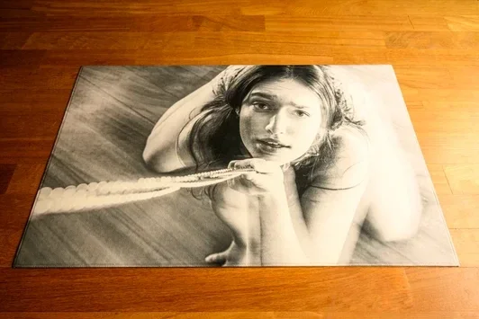 Zwart-wit vloerkleed op houten vloer met portret van knielende vrouw. De fysieke presentatie als tapijt visualiseert hoe er over een generatie wordt heengelopen.