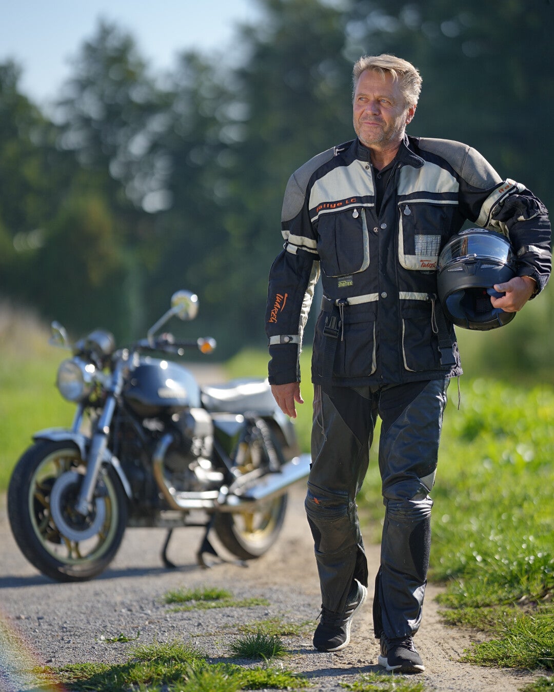 Mann in Motorradkleidung geht eine Landstraße entlang und trägt seinen Helm, im Hintergrund steht ein Motorrad.