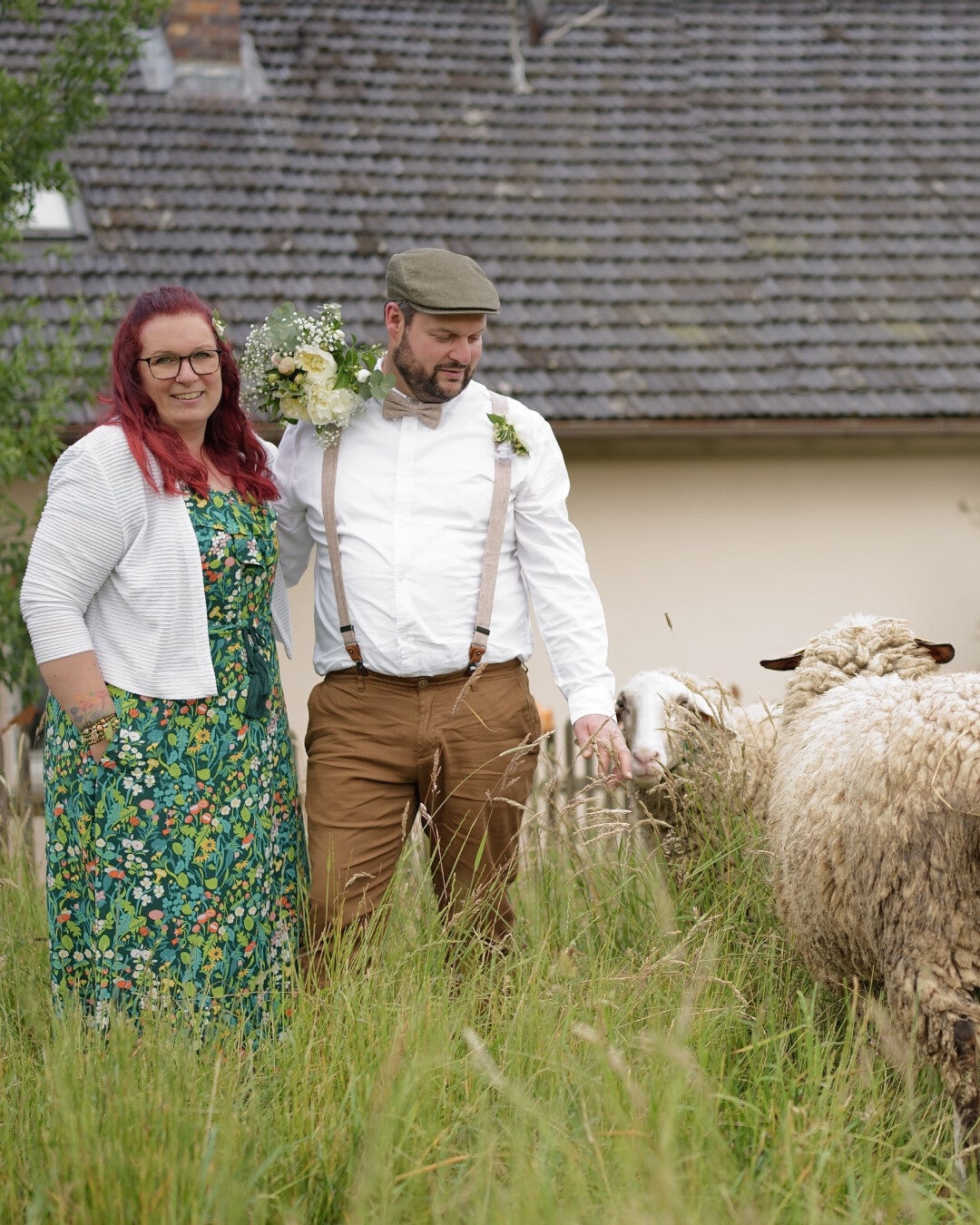 Frau und Mann stehen draußen auf einer Wiese, neben ihnen mehrere Schafe; beide tragen festliche Kleidung und lächeln.