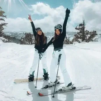 “Two women celebrating on a snowy mountain slope with skis and snowboards, representing active wellness and vitality.”