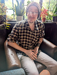 C.R.Barrett Caolan Rory Barrett sat in a leather chair illuminated by sunlight through a old wooden window with plants behind him. He sits smiling, wearing a check shirt, black beany and glasses 