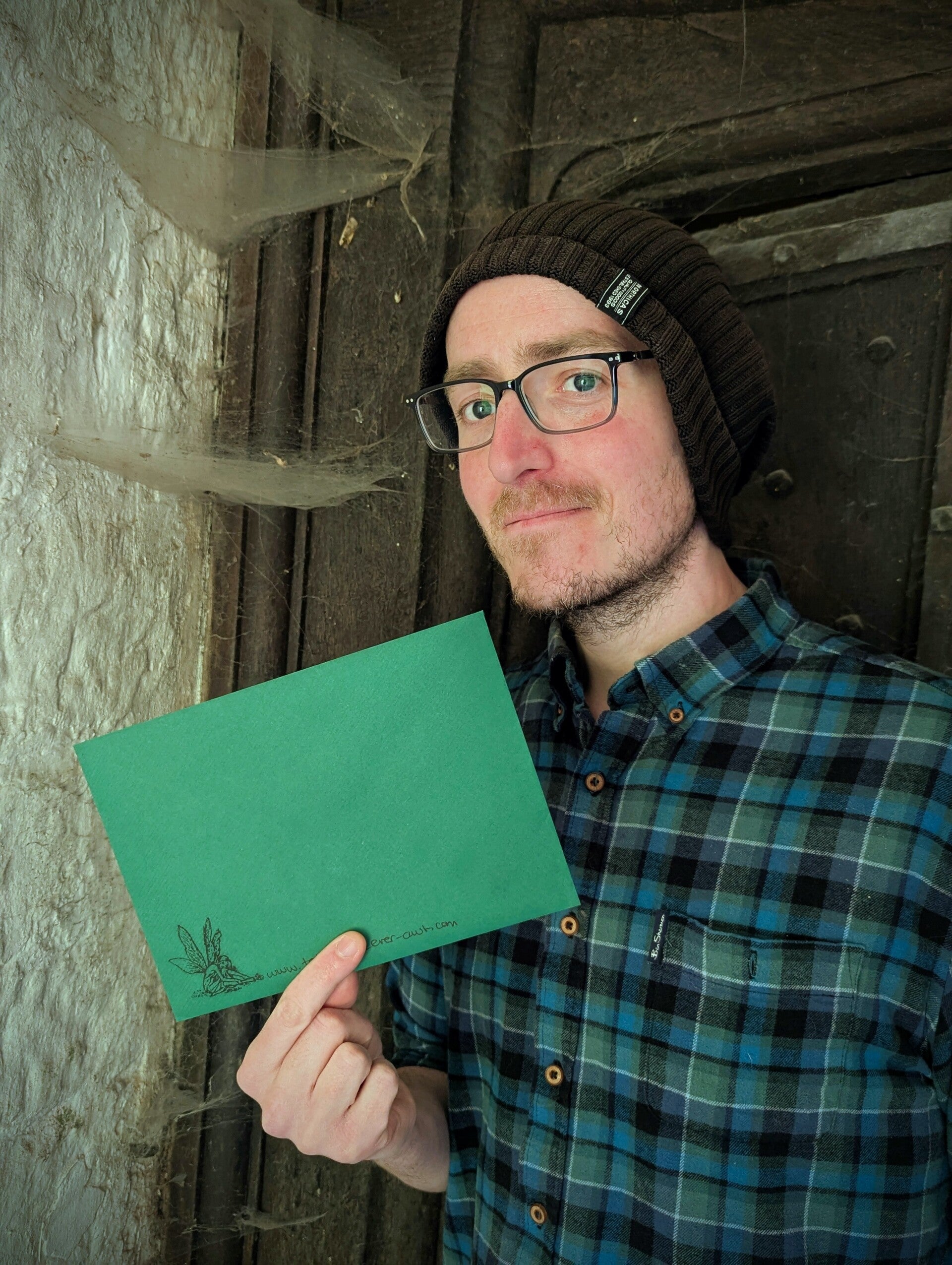 A handsome man standing in front of an ancient medieval farmhouse door holding an emerald green envelope stamped with a fairy. Tales of The Ever-Awt mail club snail mail subscription