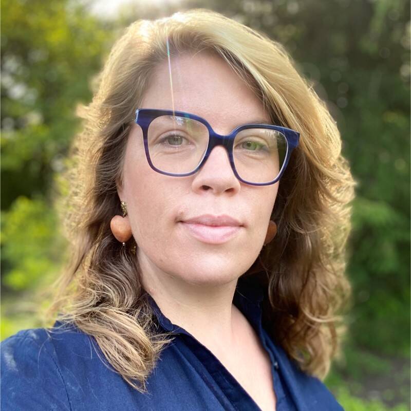 Art Historian Tascha Sciarone. Picture is of a photo of the face and shoulders of a white woman with light brown hair, blue glasses, brown earrings and a blue dress.