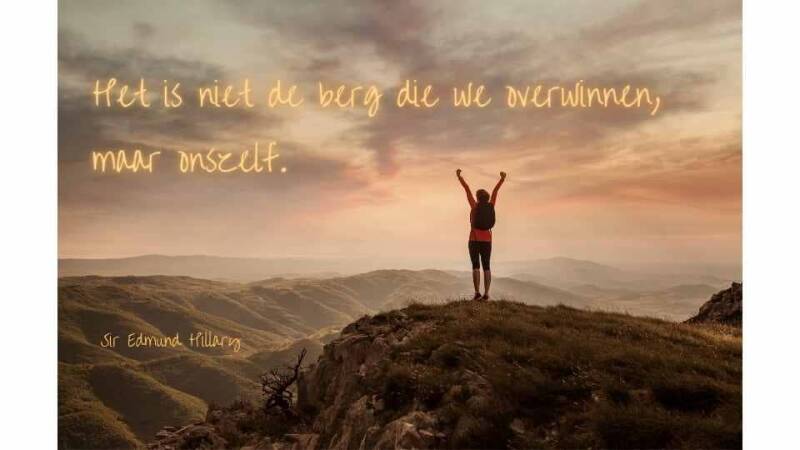 Spreuk "Het is niet de berg die we overwinnen, maar onszelf" van Sir Edmund Hillary. Op de foto staat een vrouw op een berg met  de armen in de lucht als overwinning.