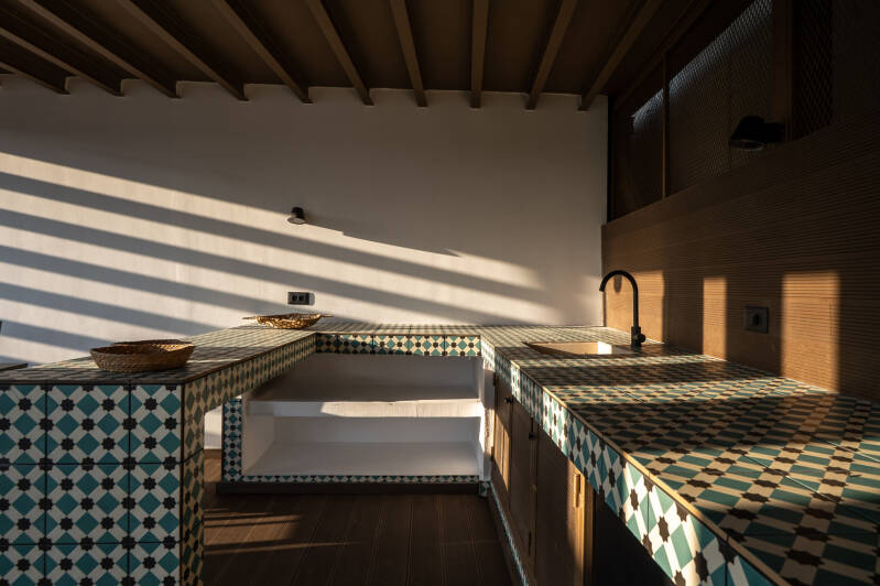 A tiled countertop kitchen with geometric patterns, shaded terrace area. Slatted shadows from the pergola roof create striped light patterns on the walls and furniture, enhancing the cozy, rustic ambiance.