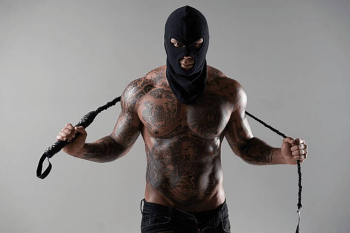 Black man with muscular body, and tattoos, wearing a black ski mask and holding a whip behind his head shows the powerful position of a dominant male in relationship dynamics