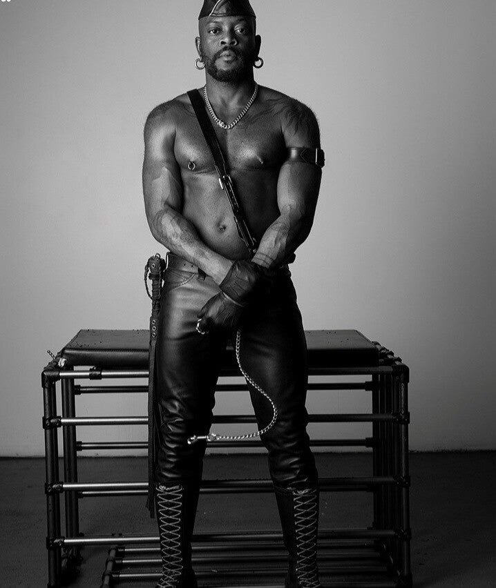 BDSMBlack and white photography of a Black male dom dressed in leather pants and harness, standing confidently against a bonding table.