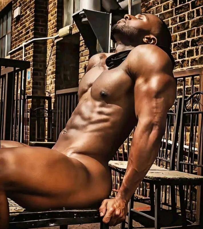 Sexy dark-skinned Black man seated outdoors, muscular physique on display, head tilted back in sunlight, near brick building and metal railings. He's exuding confidence in his body as he poses peacefully in the nude, Elevating the eroticism