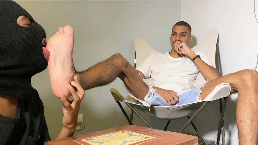 Black man sitting in a chair as a white man wearing a mask kneels at his feet in a ritualistic foot worship session.
