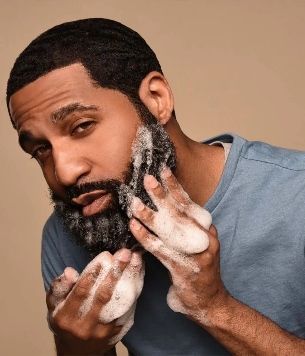 Older handsome Black male with low-cut, wavy hair grooming his beard while looking in a mirror, showcasing a well-styled appearance and confident masculinity