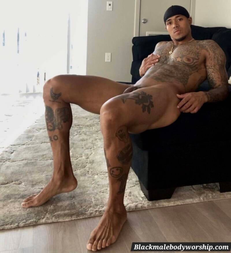 Shirtless tattooed man in black bandana poses erotically on a black couch, showing muscular body and well-groomed, long toes for foot fetishism and Black male feet appreciation 