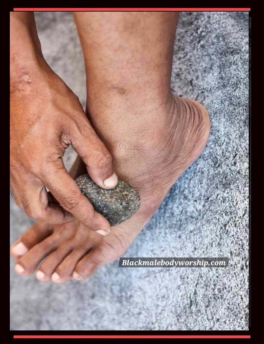 Up-close top view of a barefoot Black man using a pumice stone on the side of his foot, engaging in proper foot care to remove dry, callused skin.