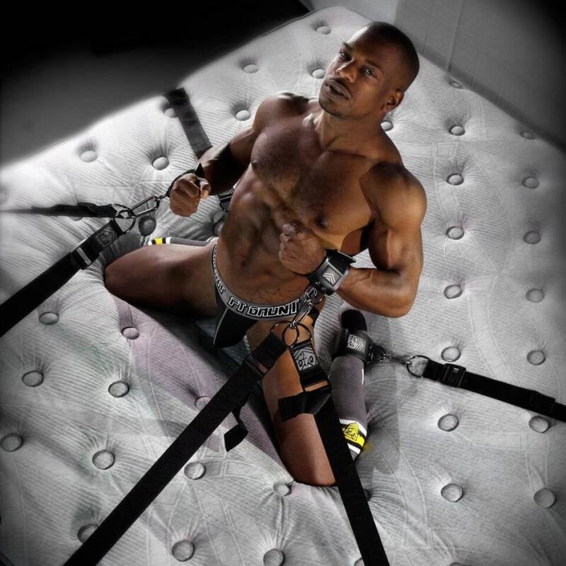 A powerful Black man in a harnessed restraint system on a mattress, representing masculine exploration, intimacy, trust, and physical control. Wearing a jockstrap, he embodies strength, vulnerability, and intentional connection in a private moment.