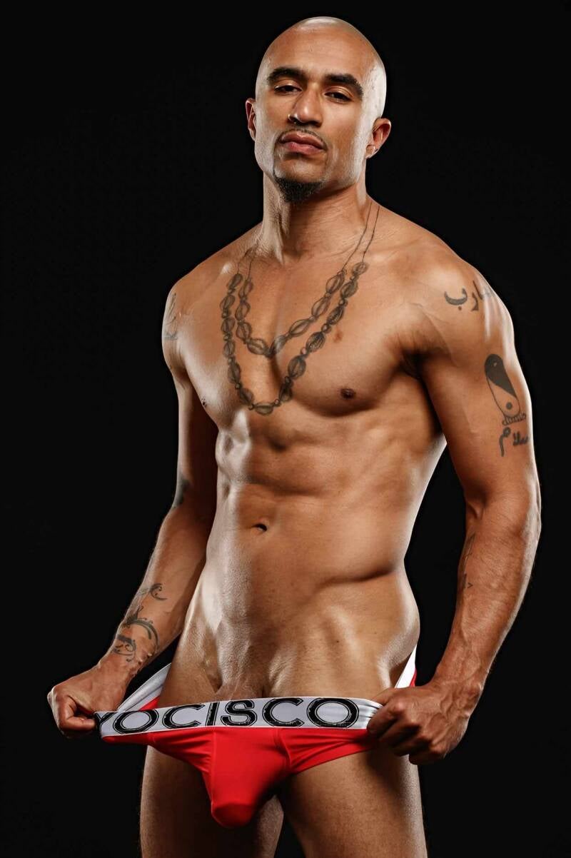 King Noire, a light-skinned, bald man, stands shirtless against a black backdrop, his muscular chest adorned with a long beaded necklace. Tattoos cover his arms and shoulders as he grips the waistband of red YOCISCO briefs, displaying the white brand name