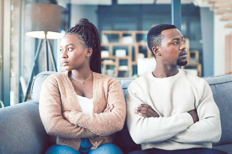 Upset African American couple sitting on a couch with arms crossed, showing signs of relationship problems and emotional disconnect.