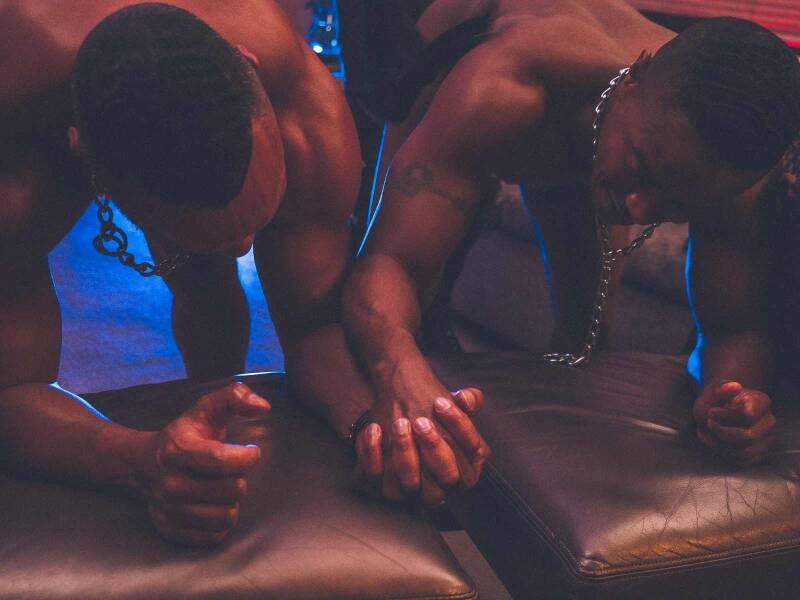 "Gay Black submissive men honoring power exchange while wearing choke collars in a consensual BDSM ritual, expressing emotional surrender, deep trust, and erotic intimacy."