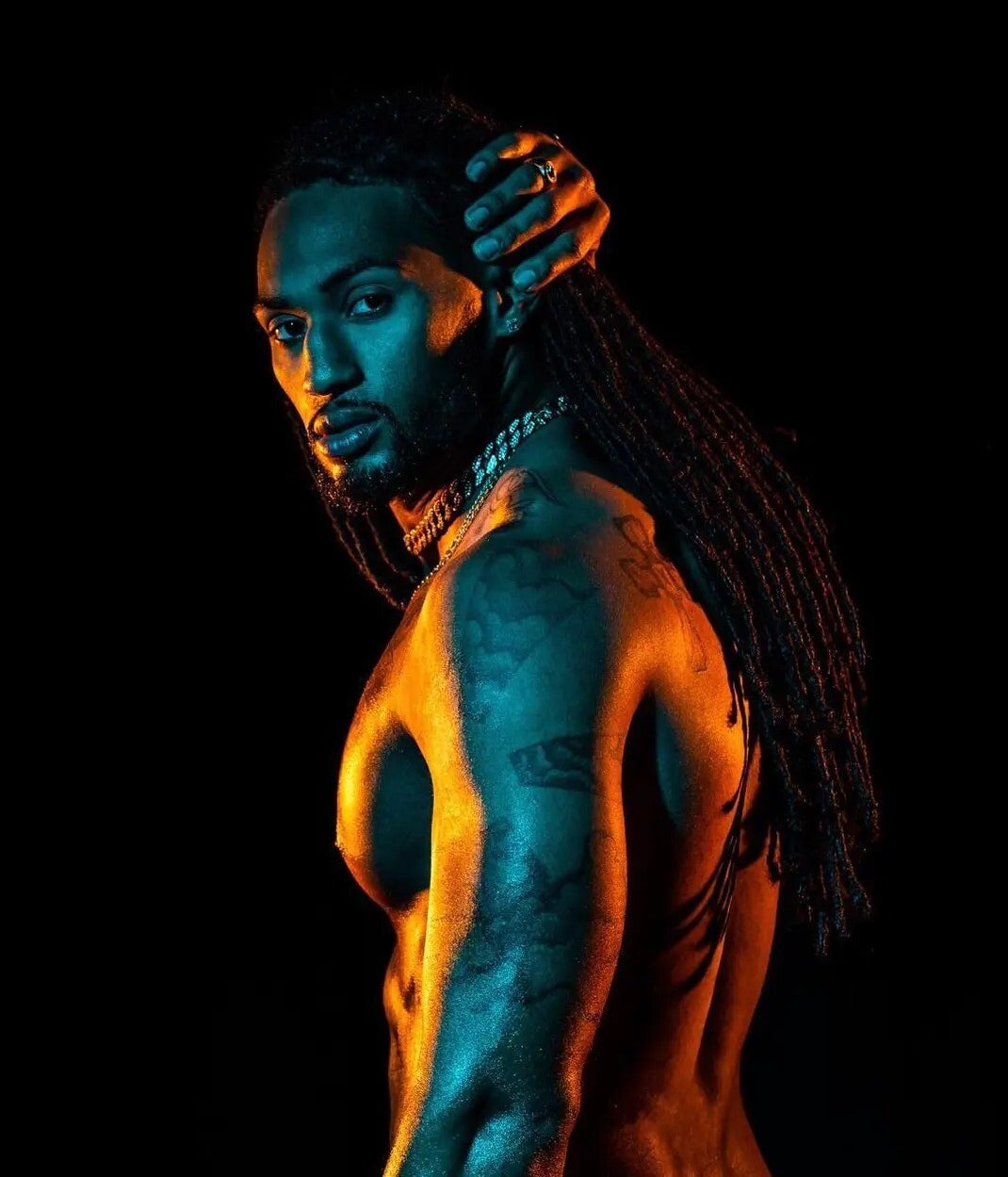Alluring Sexy Black male shirtless, with tattoos, and dreads posing his muscular physique from a side up-close angle against a dark background, with a fluorescent lighting across his face–making him look mysterious, confident, attractive, and masculine