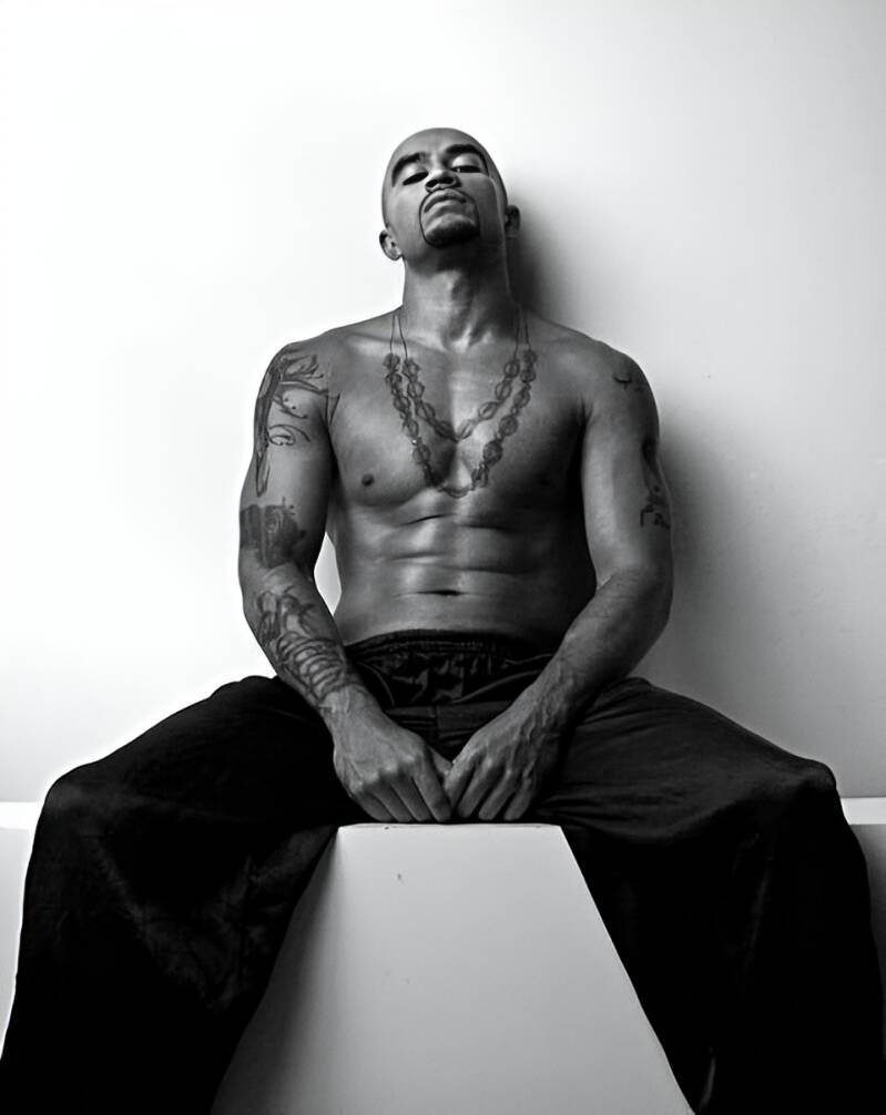 Black and white portrait of muscular, tattooed man with necklace seated against white wall, reflecting on failed relationships, self-help quick fixes, and the need for sexual compatibility and honesty.