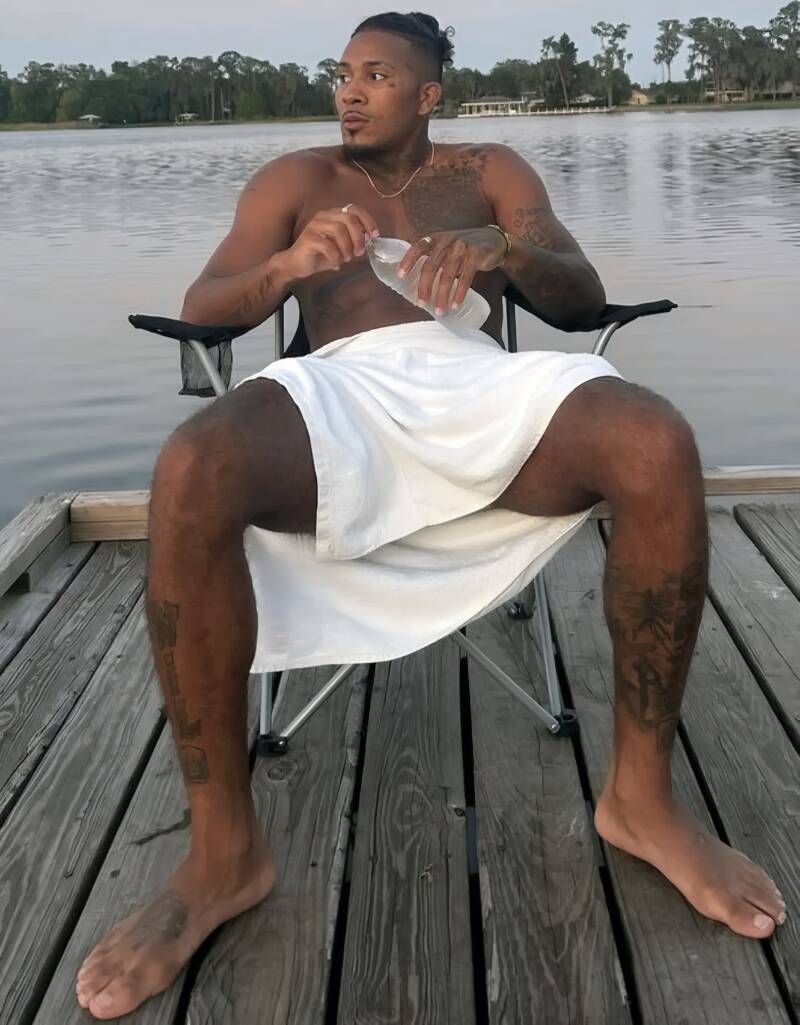 shirtless, muscular, light-skinned Black man with tattoos sits on a black folding chair on a wooden dock, with his huge bare feet on display to be worshipped and cherished.