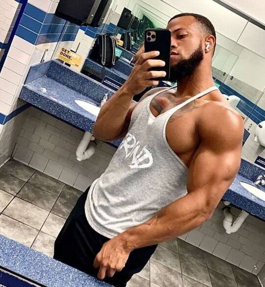 Maurice Holmes fitness coach, life coach, and erotic wellness coach looking masculine and strong—posing In gym bathroom in grey tank top show casing masculine physique and huge bisceps.