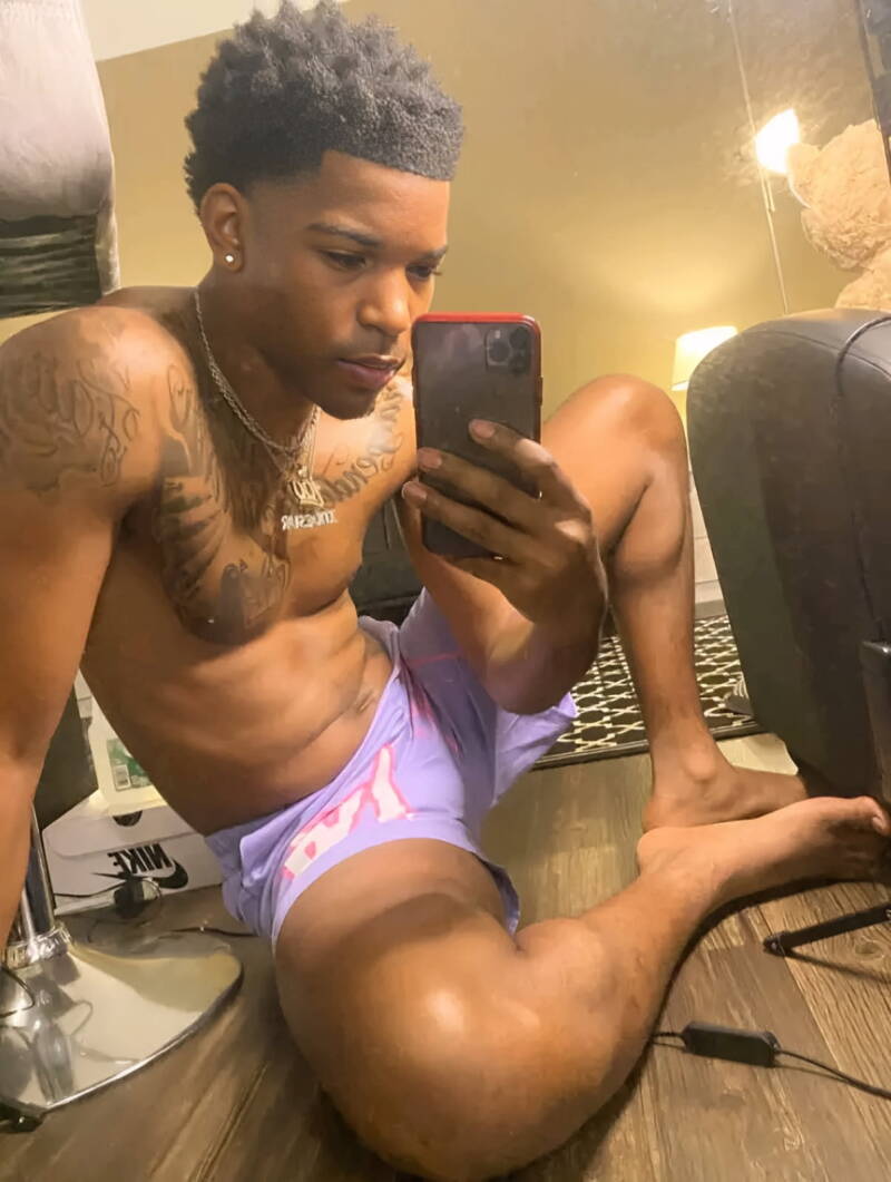 Tattooed Black man taking a selfie on the floor, side angle view showing clean, athletic feet with strong arches and defined toes, wearing lavender shorts.