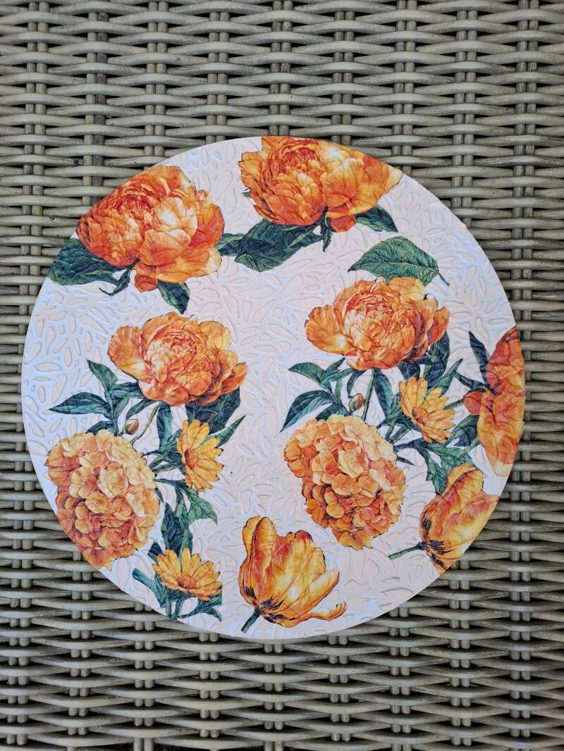 Handgemaakte muurcirkel van 30 cm met reliëfpasta en decoupage van oranje bloemen op een lichtgekleurde achtergrond met subtiele structuur.