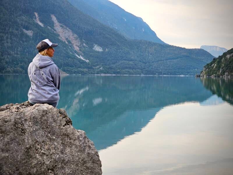 Ruhiger Bergsee mit kristallklarem Wasser: Eine Frau sitzt auf einem Felsen am Ufer und genießt die friedliche Aussicht auf die spiegelnde Wasseroberfläche und die umliegenden Berge.