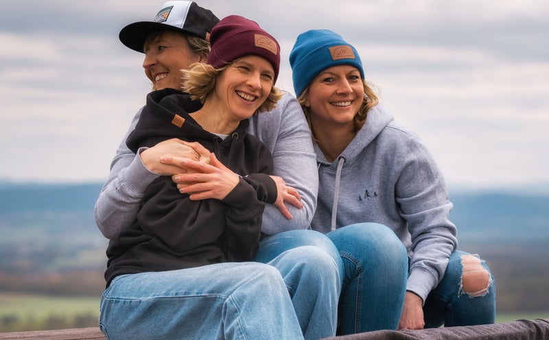 Drei Personen sitzen auf einer Holzbank auf einem Berg. Sie tragen Hoodies und farbige Beanies, die ein urbanes Outdoor-Feeling vermitteln. Auf der Bank liegt eine schwarze Decke mit einem stilisierten Seil- und Karabiner-Motiv sowie einem Schriftzug.