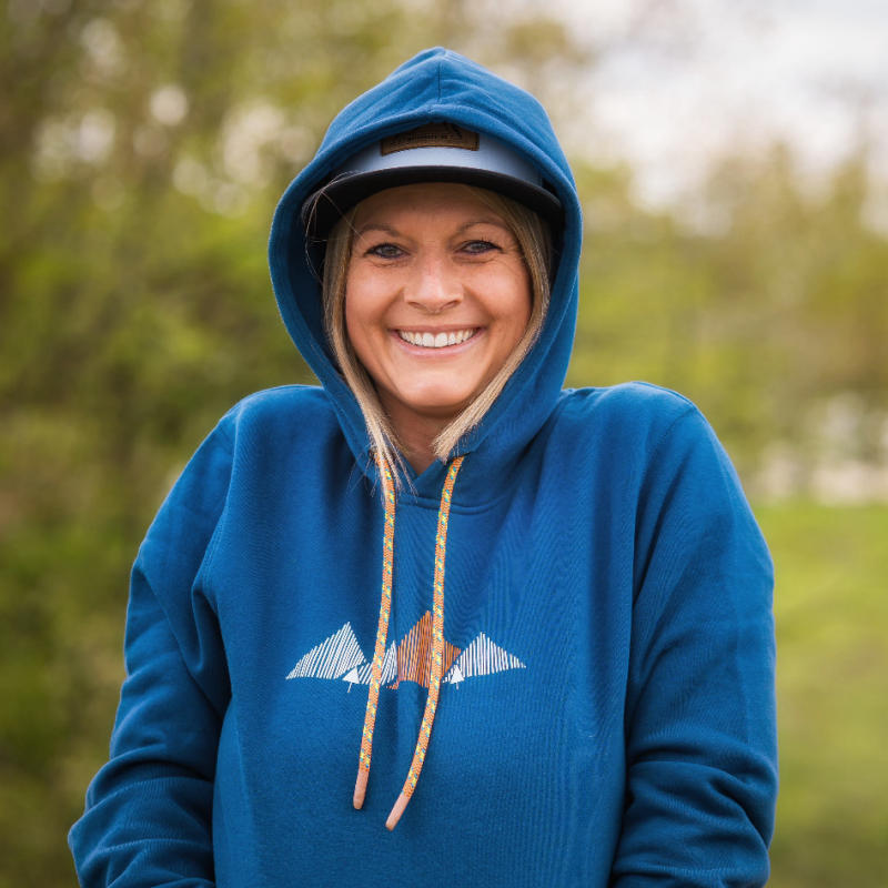 Eine blonde Frau trägt einen blauen Hoodie mit einem stilisierten Bergmotiv auf der Vorderseite und farblich passenden Kordeln, die Outdoor- und Abenteuerstimmung vermitteln. Der Hintergrund zeigt eine natürliche, grüne Umgebung