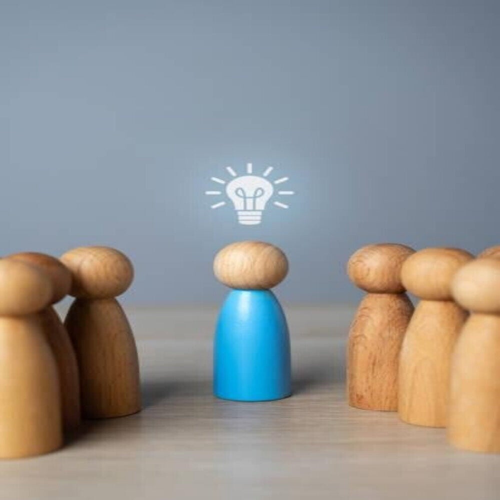 Holzfiguren, eine blaue in der Mitte mit einer gezeichneten Glühbirne darüber, umgeben von mehreren naturfarbenen Holzfiguren, im Kontext mit MediumSciences - Institut.