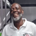 A smiling man in a white "KEL'S AUTO SERVICE" polo shirt and jeans stands in front of shelves stocked with automotive supplies, including oil containers, car polish, paint cans, and windshield wiper blades.