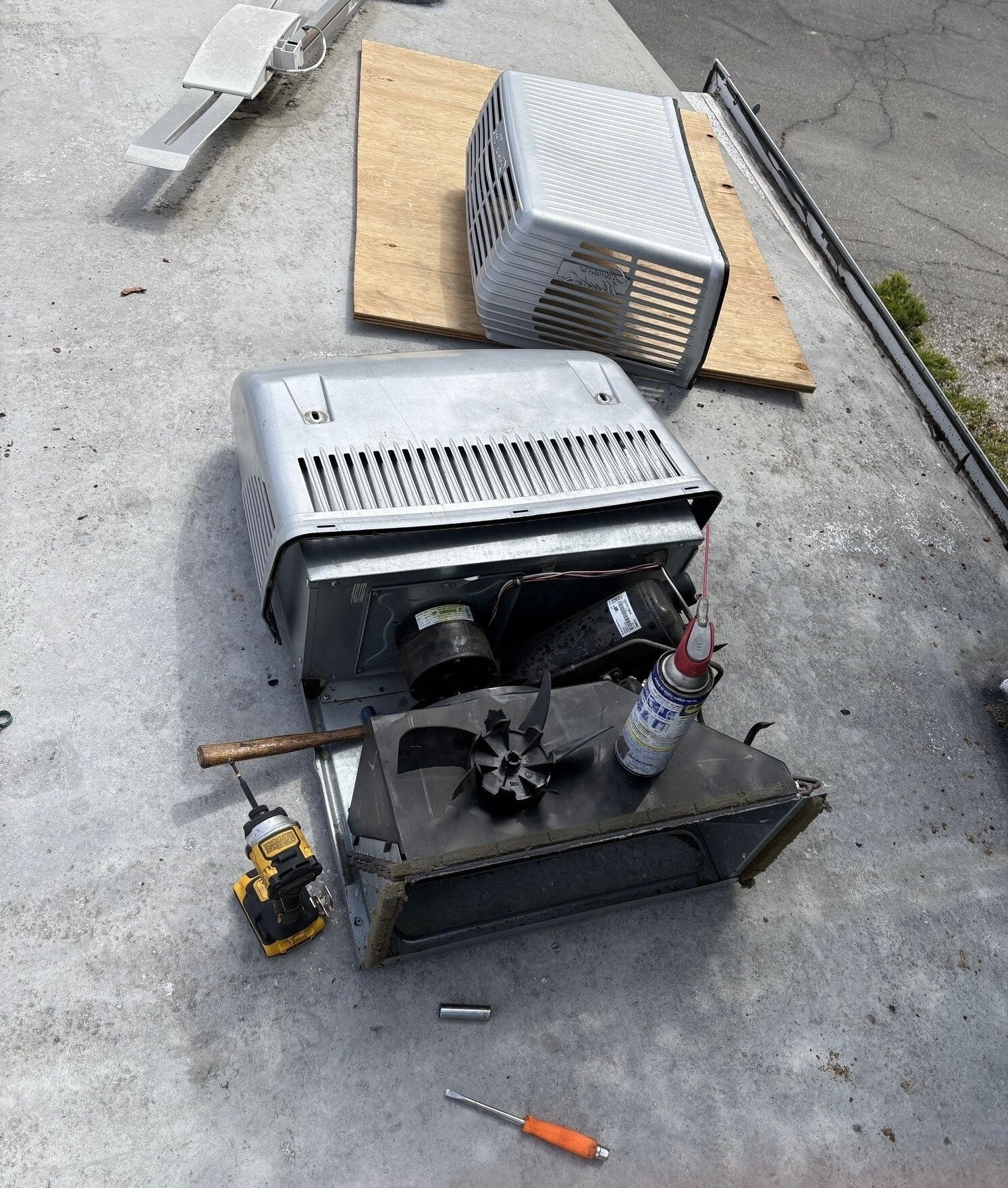 RV rooftop air conditioner with the cover removed showing the newly replaced blower fan during DIY repair on concrete driveway, tools including cordless drill, screwdriver, and cleaning spray nearby