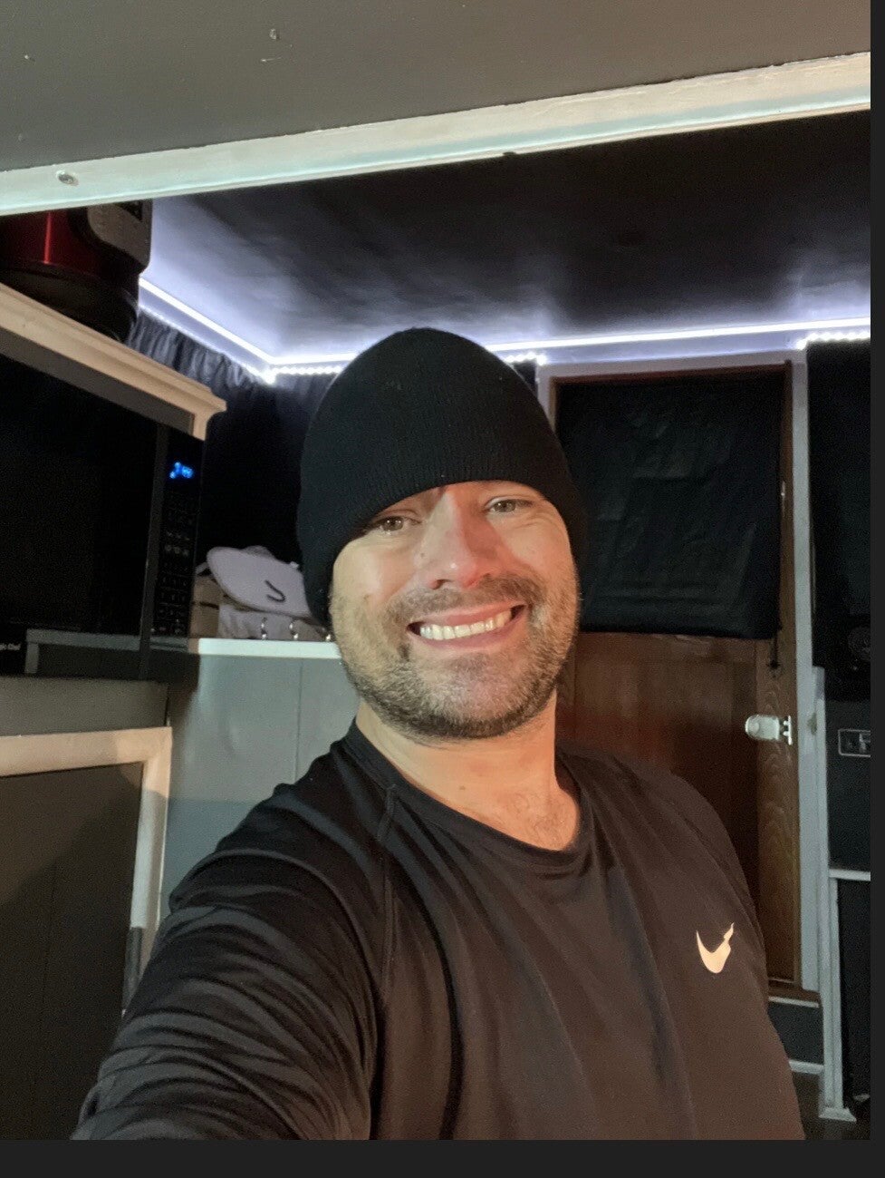 Smiling man with a short beard, wearing a black beanie and black long-sleeve shirt, taking a selfie inside the kitchen of an RV or camper van. LED lighting runs along the ceiling with a microwave and cabinets visible in the background
