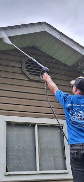 Worker pressure washing brown house siding near roof eave.