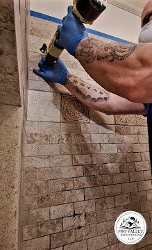 Detail view of a contractor's tattooed arm and blue gloved hand using a professional caulking gun to repair grout lines on a travertine stone shower wall by Pro Valley Renovations LLC.