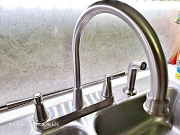Detail shot of a high-arc brushed nickel kitchen faucet and side sprayer installed on a stainless steel double sink with a marble-pattern backsplash.