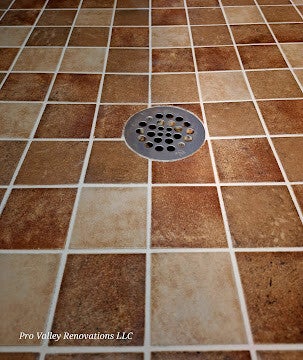View of a stainless steel circular shower floor drain set into a mosaic of square rustic, textured brown and beige stone tiles.