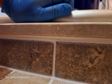 Macro shot of a worker's blue-gloved hand performing delicate detailing work along a tile edge, with watermark text partially visible.
