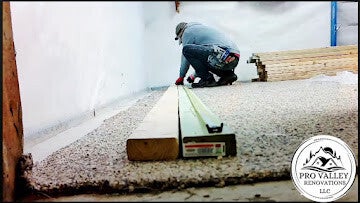 Worker leveling ground surface using straightedge and wooden beam near exterior wall.