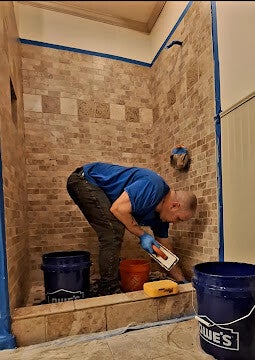 A professional contractor crouching and using a float to apply grout to a large travertine stone block shower wall with Lowe's buckets nearby.