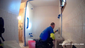 A painter in a blue shirt on hands and knees preparing to paint the walls of a small bathroom with a sloped ceiling, showing blue painter's tape on all edges.