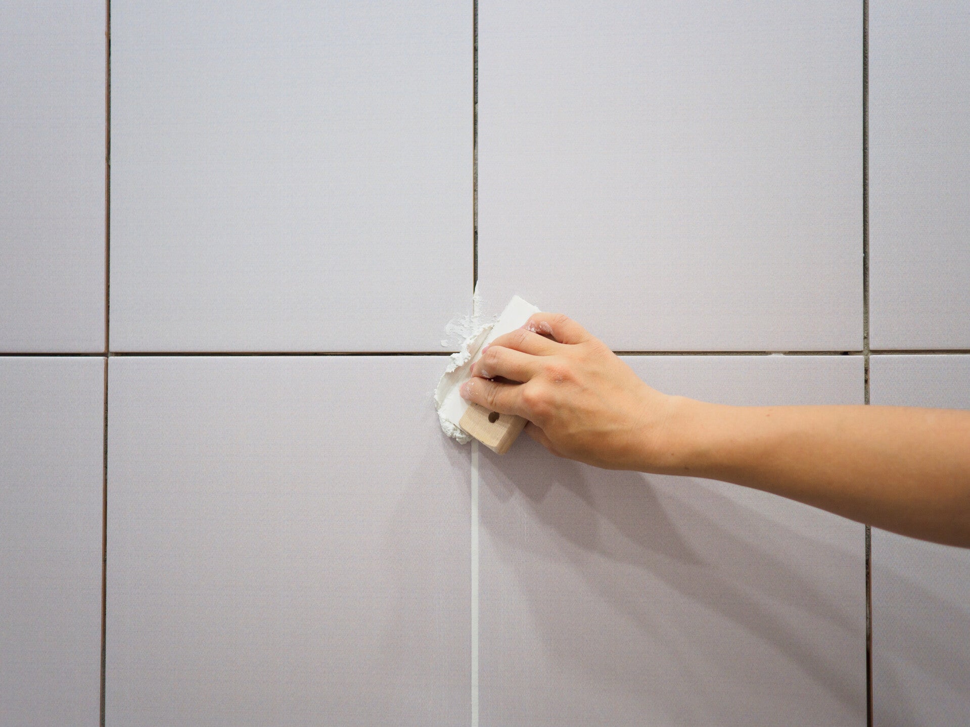 Professional grout repair technique showing a contractor using a clean grout sponge to wipe away excess wet grout from light grey wall tiles.