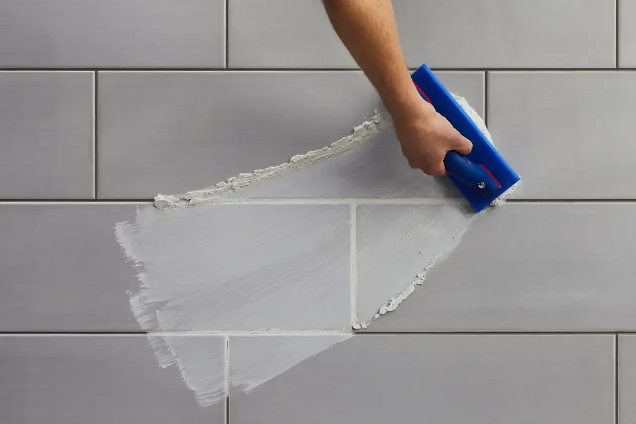 An overhead view of a contractor's hand using a blue grout float to apply light grey grout to horizontal subway wall tiles.