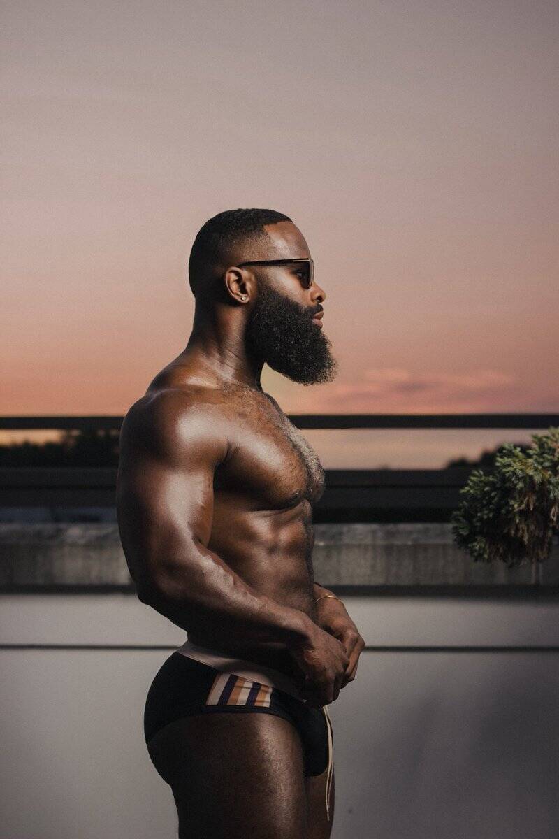 Black fitness model standing sideways showing off side profile revealing broad chest, big pecks, six pack, rounded butt, and toned masculine thighs in front thick muscular thighs in front of a sunset.