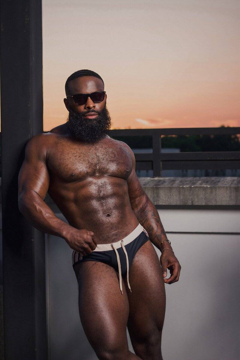 Black male model showing off stunning body while posing in underwear up against a wall and a beautiful outside background 