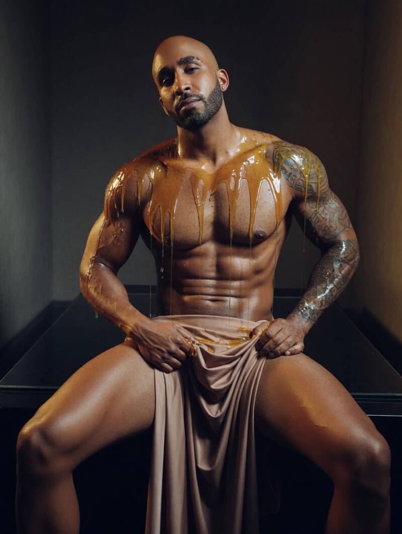Full-shot of an athletic, shirtless Black man with a defined six-pack and tattoos, seated and holding a light-colored cloth over his lap. Golden liquid drips down his shoulders and chest against a dark studio background.